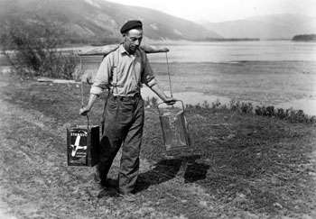 Claude carries two gas cans of water on a yoke up the riverbank at Forty Mile.